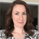 A woman with shoulder-length brown hair, wearing a patterned top, stands indoors at home and smiles at the camera.